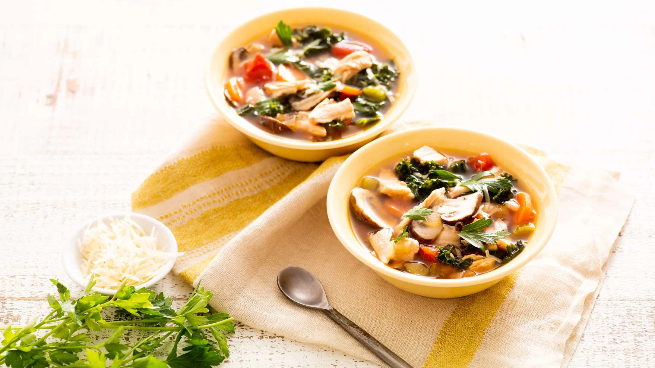 Two bowls of hearty vegetable soup with chicken, mushrooms and greens on a yellow cloth with fresh herbs nearby.