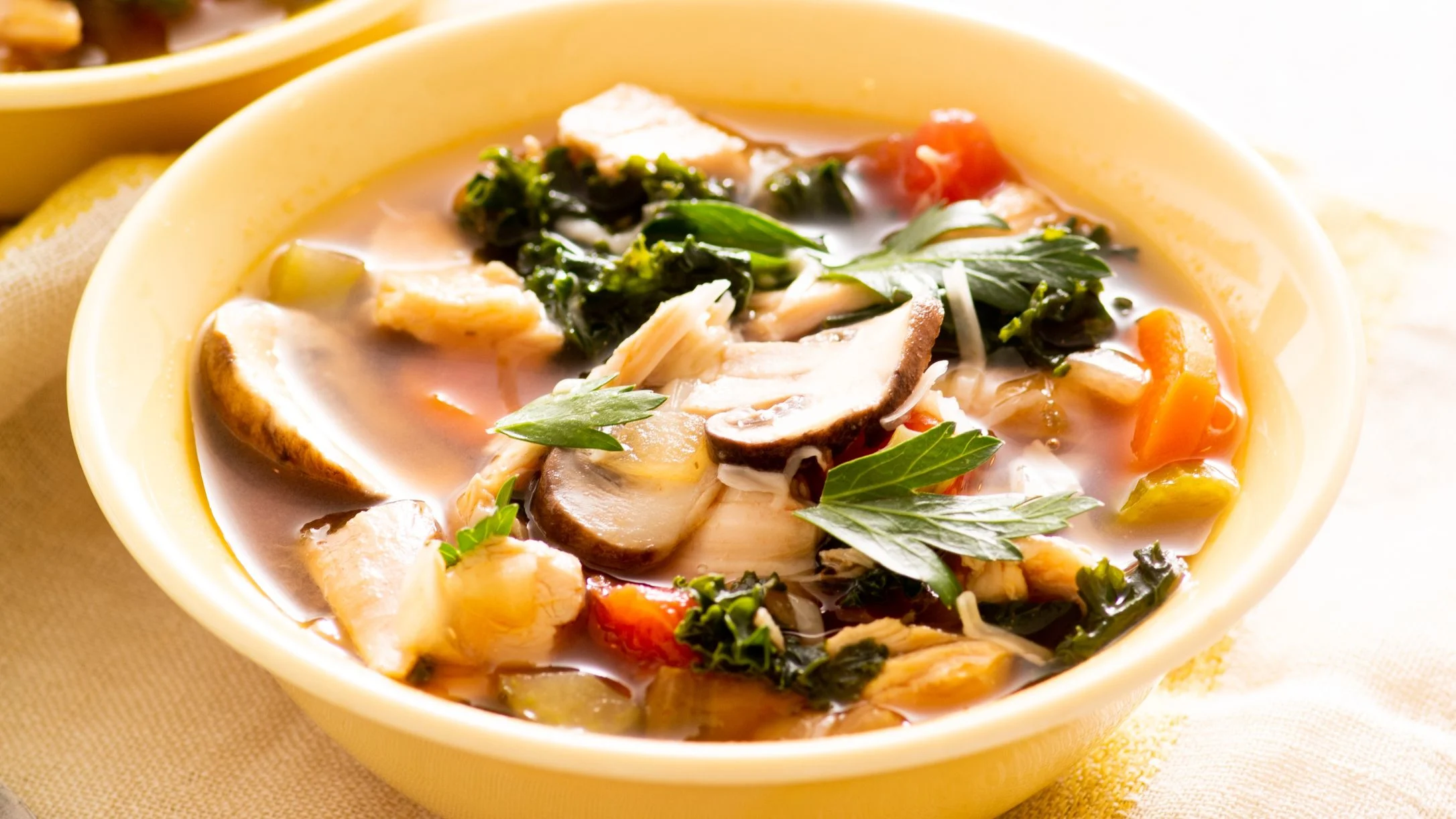 Hearty vegetable soup in yellow bowl with mushrooms, kale, chicken, and herbs in clear broth.