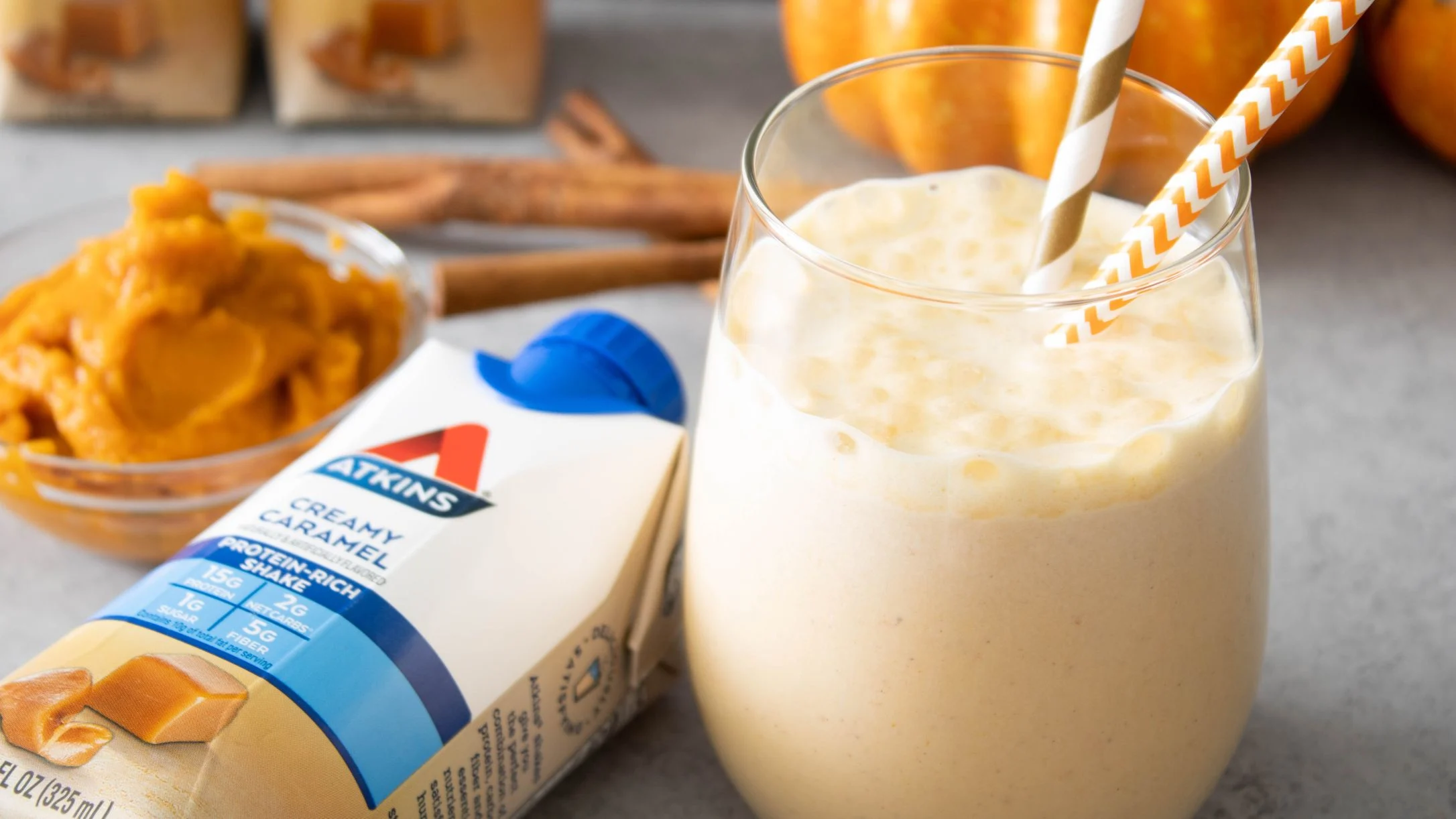 Creamy pumpkin smoothie with paper straws next to Atkins Creamy Caramel shake and a bowl of pumpkin puree.