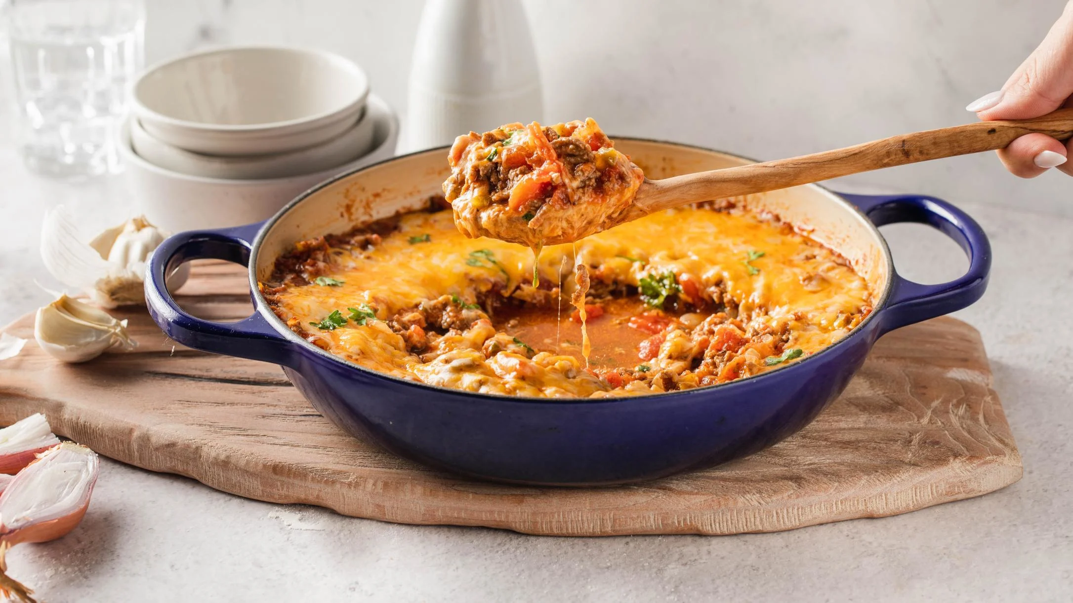 Wooden spoon lifting cheesy meat casserole from blue baking dish, with melted cheese stretching from the spoonful.