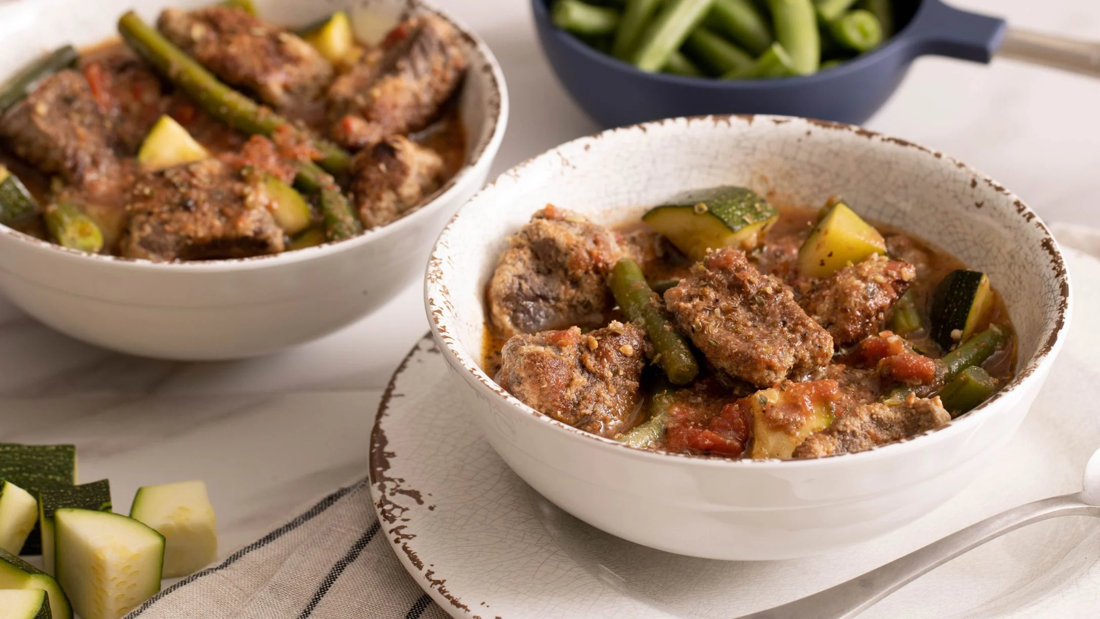 Two white bowls of meat stew with zucchini and green beans, with fresh vegetables and a side dish nearby.