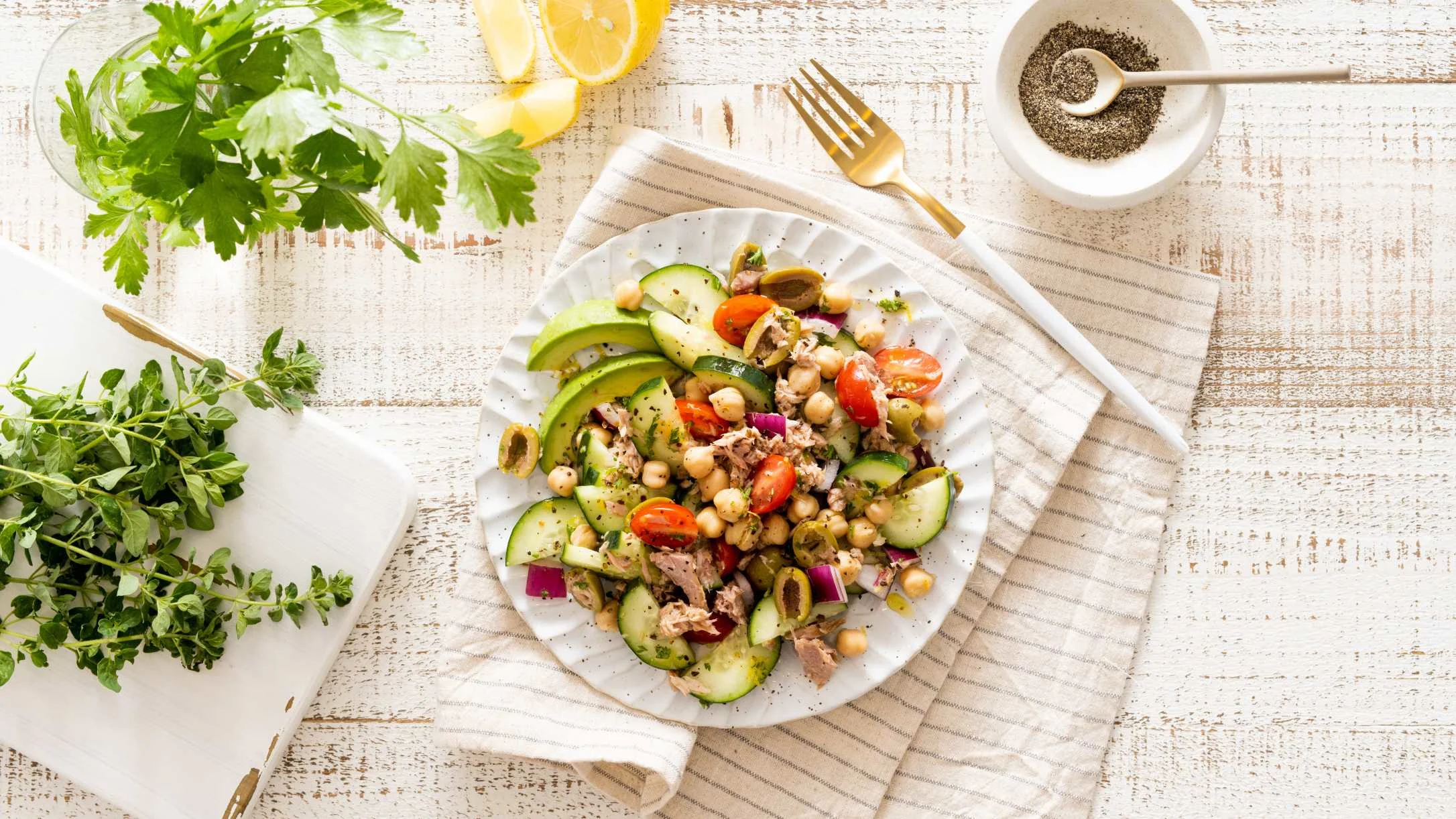 Fresh Mediterranean salad with chickpeas, avocado, cucumber, and tomatoes on white plate, surrounded by herbs and spices.