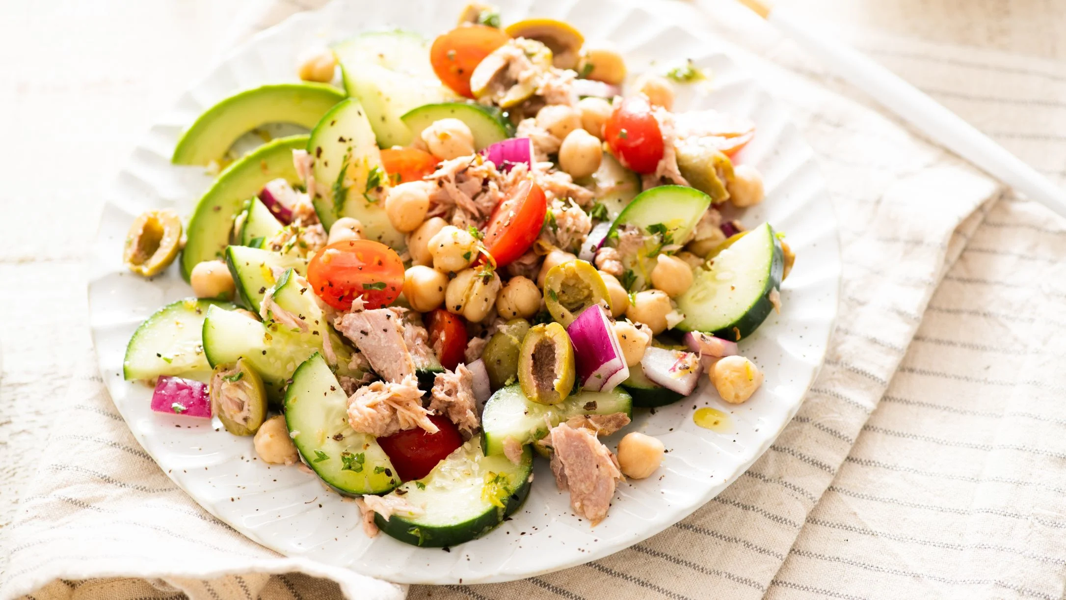 Fresh Mediterranean salad with tuna, chickpeas, cucumber, avocado, cherry tomatoes, and red onion on a white plate.