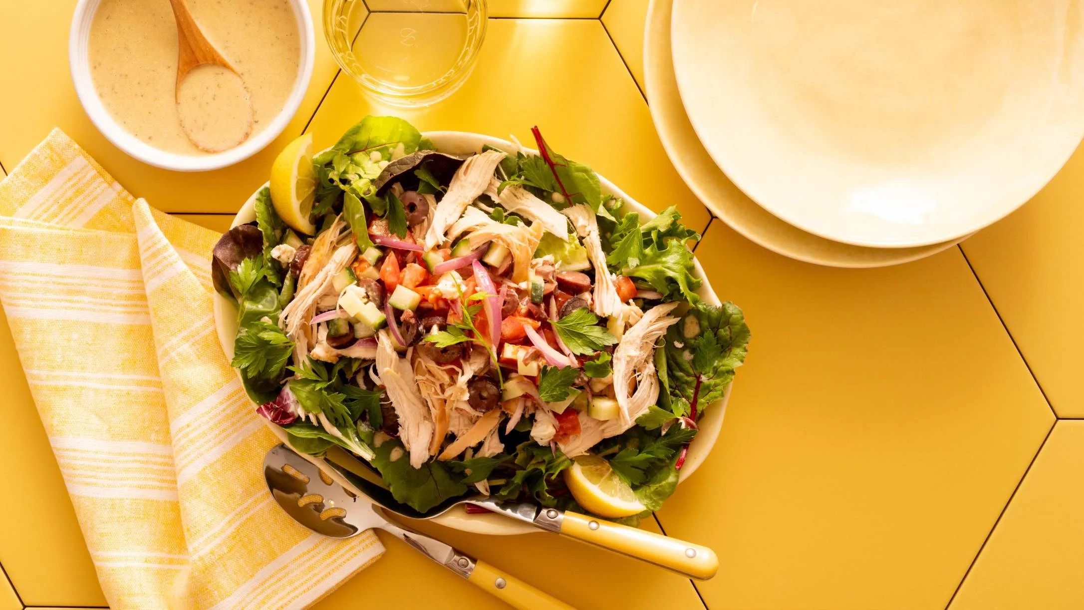 Chicken salad with fresh vegetables on yellow table, served with dressing, empty plate, and yellow napkin.