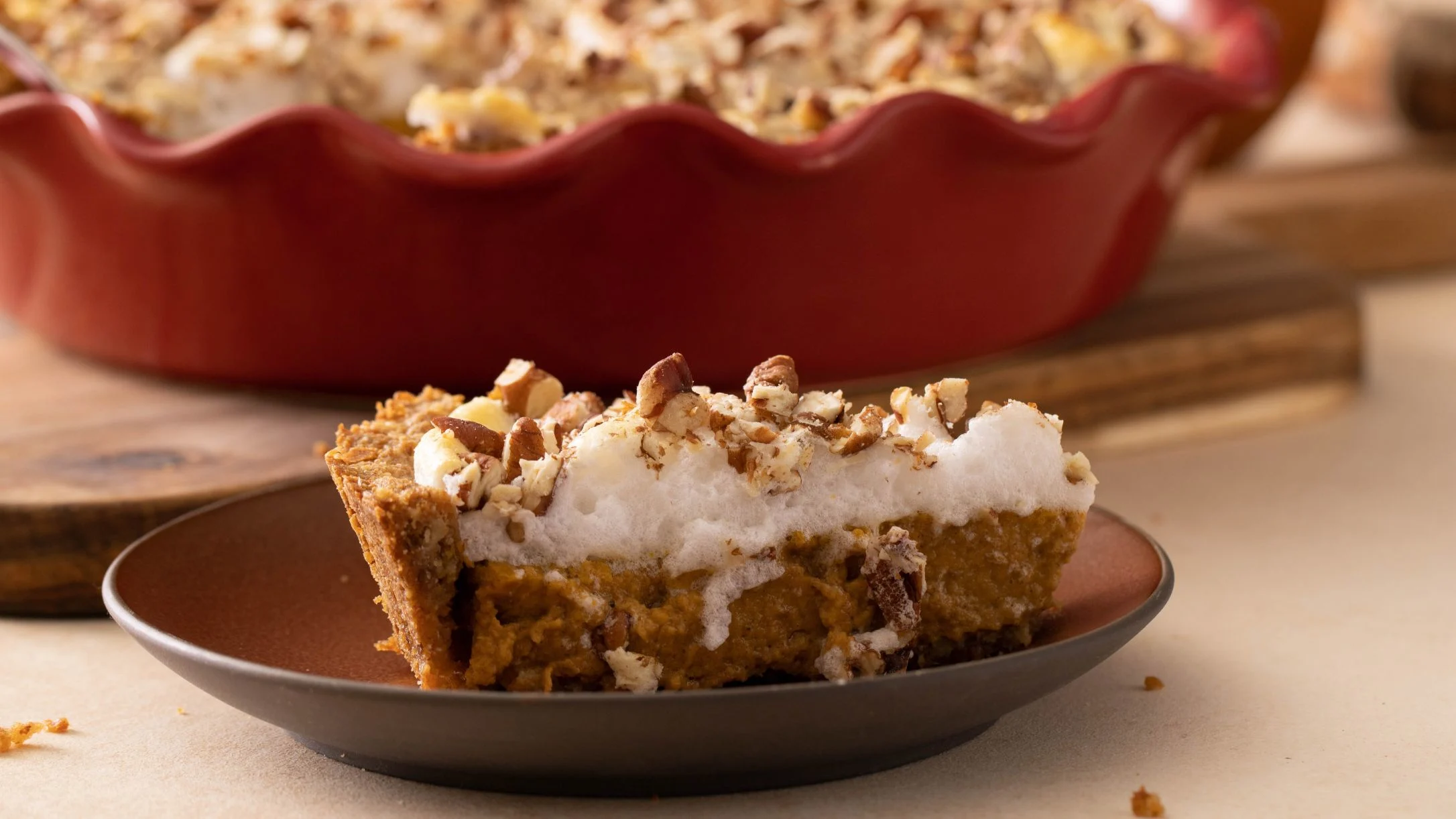 Slice of pumpkin pie with meringue and chopped nuts on a brown plate, with red pie dish in background.