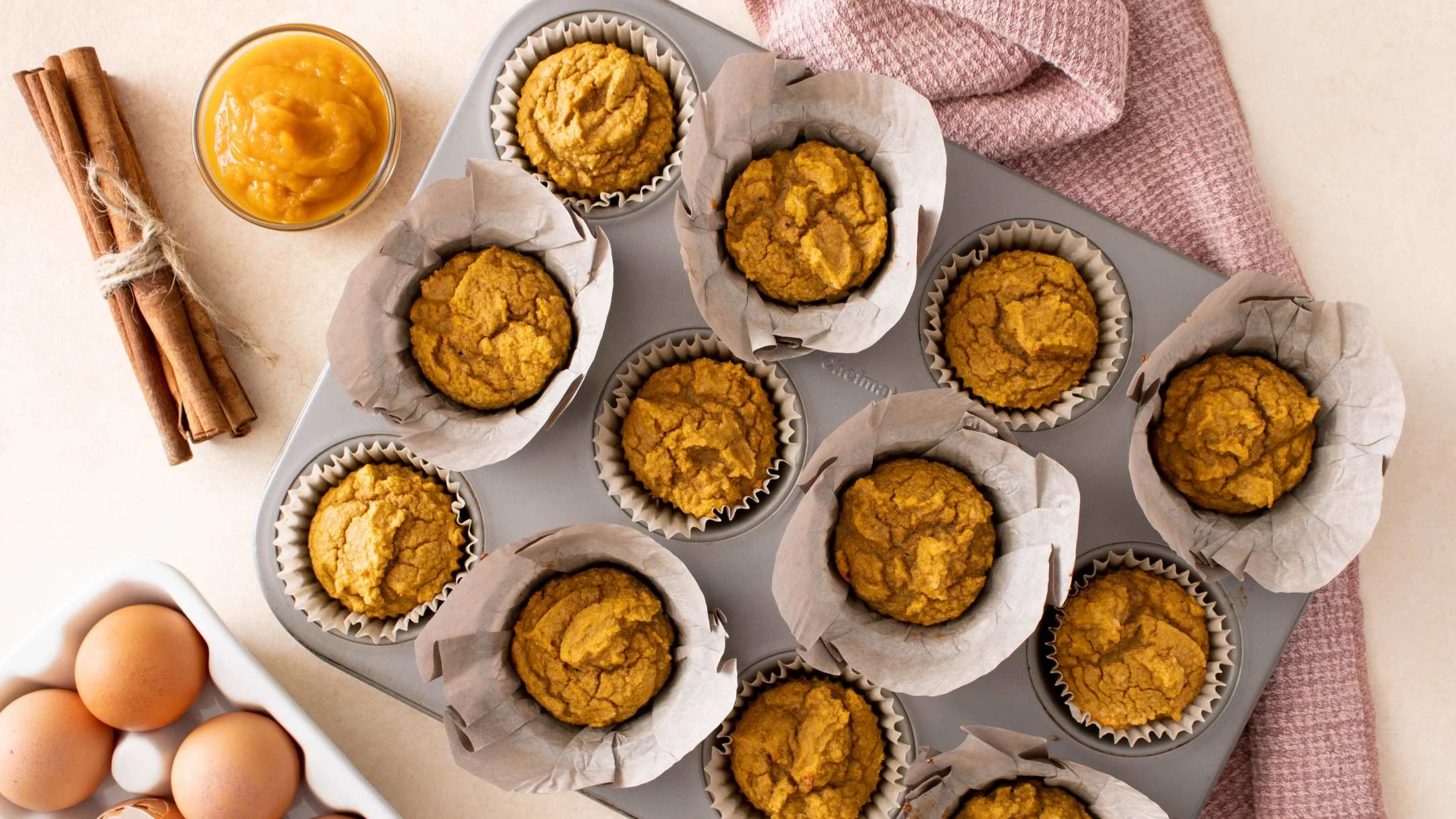 Freshly baked pumpkin muffins in a tin with pumpkin puree, cinnamon sticks, and eggs on the side.