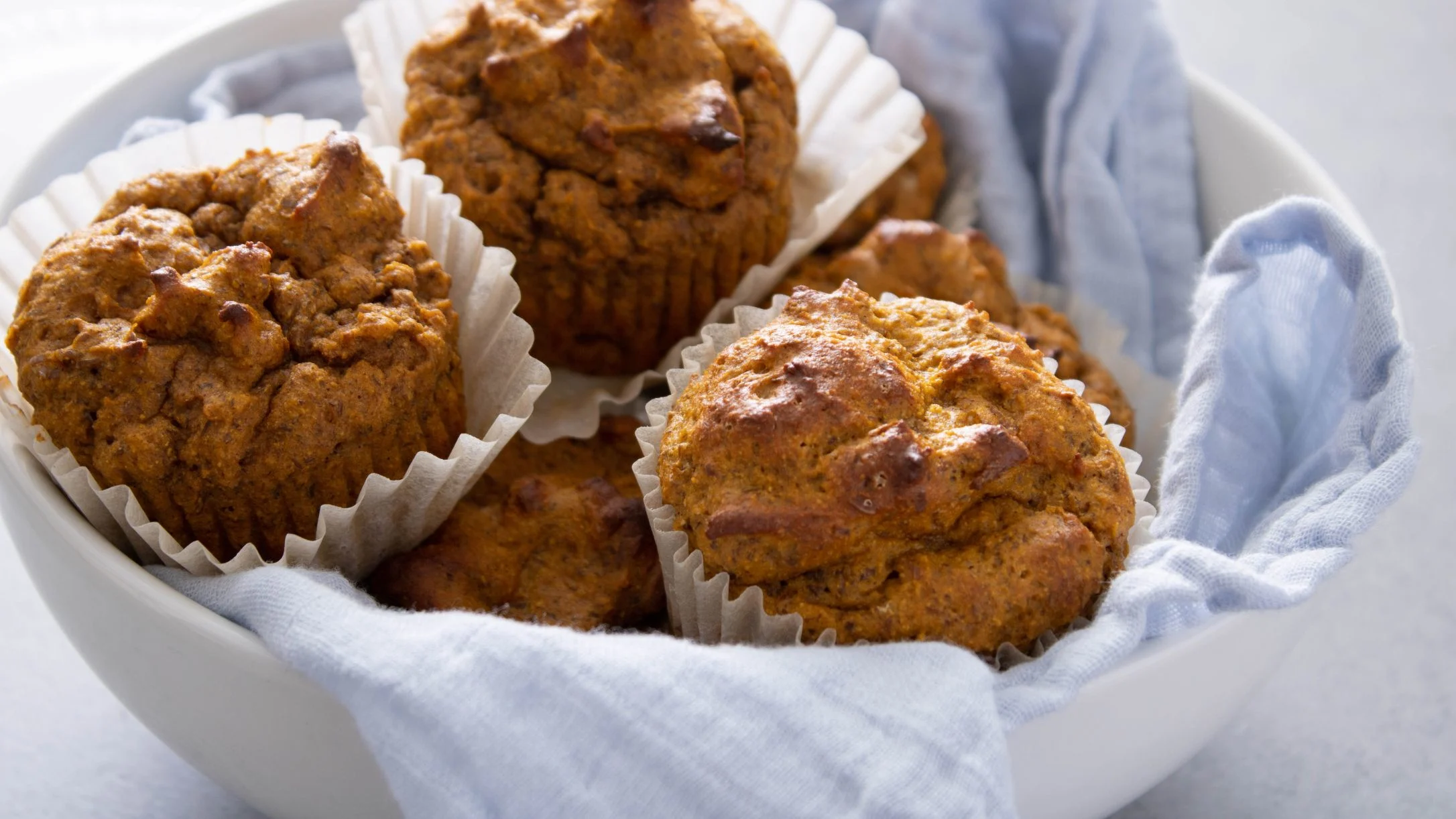 Freshly baked pumpkin muffins in paper liners, served in a white bowl lined with light blue cloth.