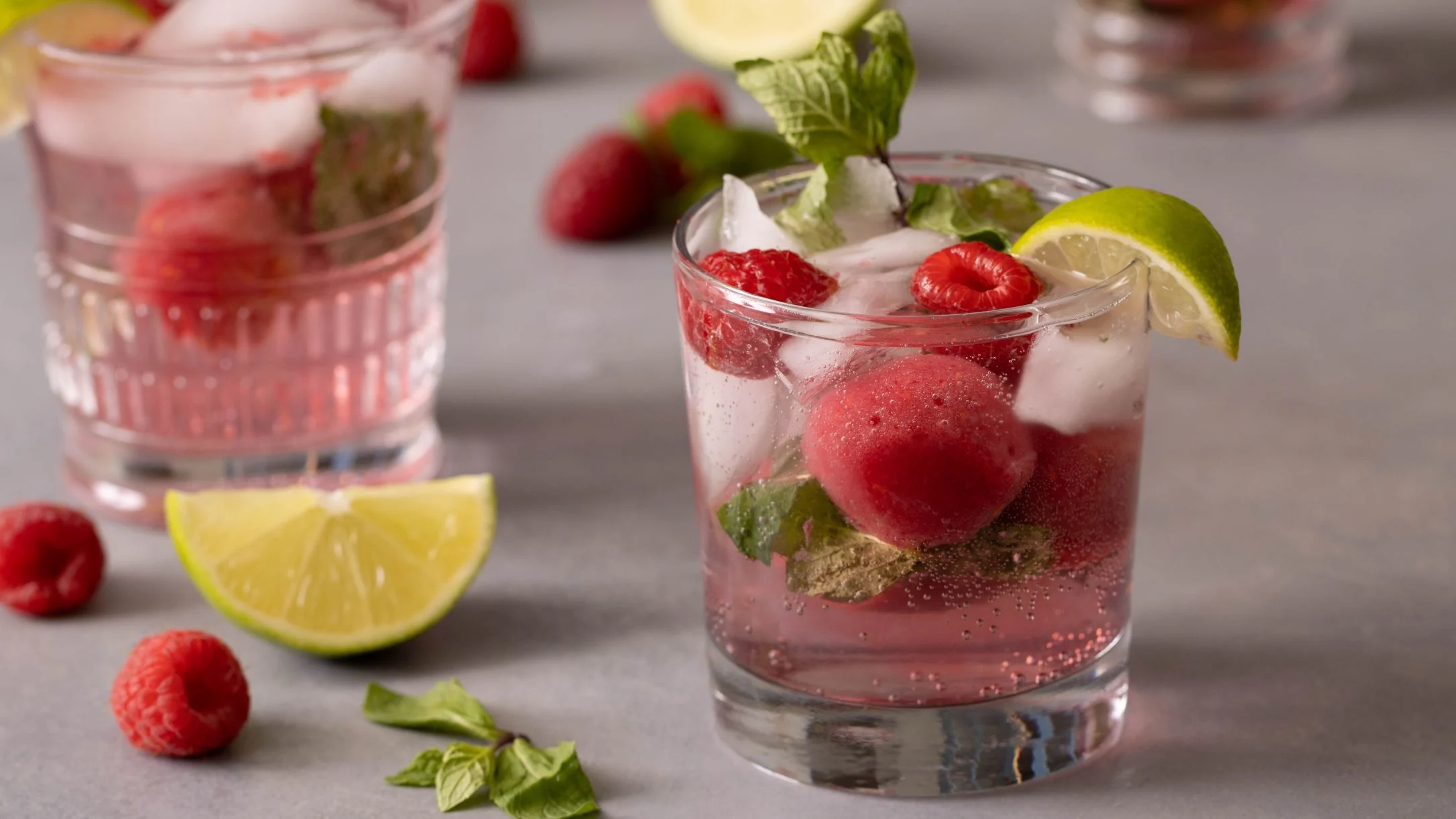 Refreshing raspberry cocktails in crystal glasses garnished with lime slices, mint leaves, and fresh berries on ice.