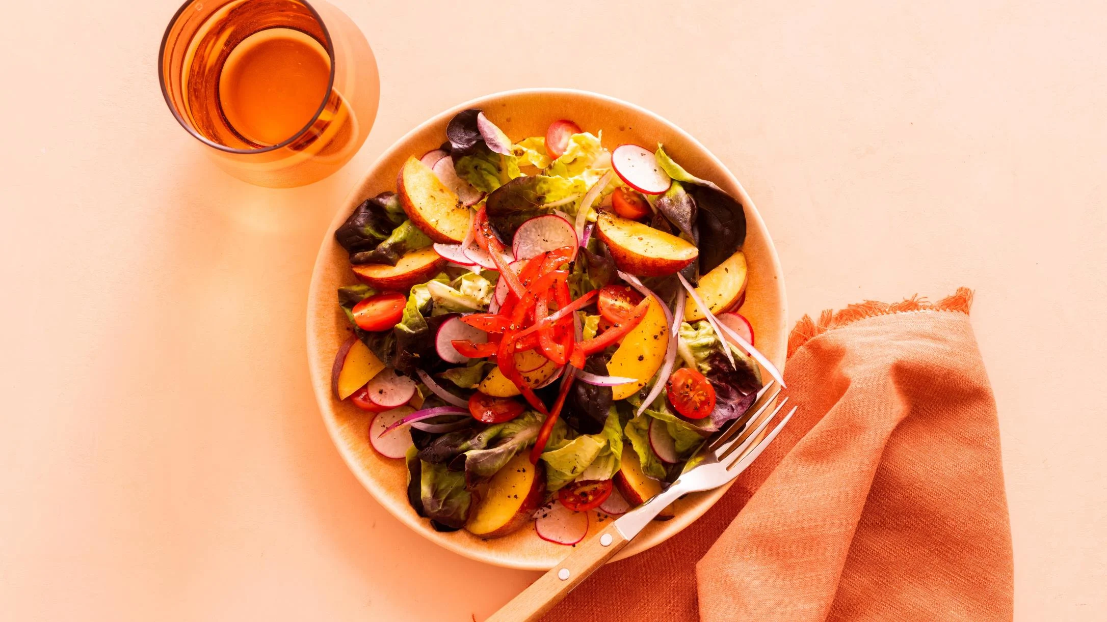Fresh garden salad with peach slices, radishes, tomatoes and greens in a beige bowl, served with orange drink.