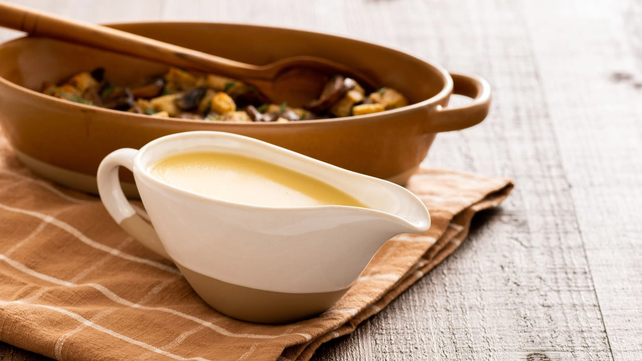 White gravy boat with creamy sauce beside brown ceramic dish containing mushroom mixture on striped cloth napkin.