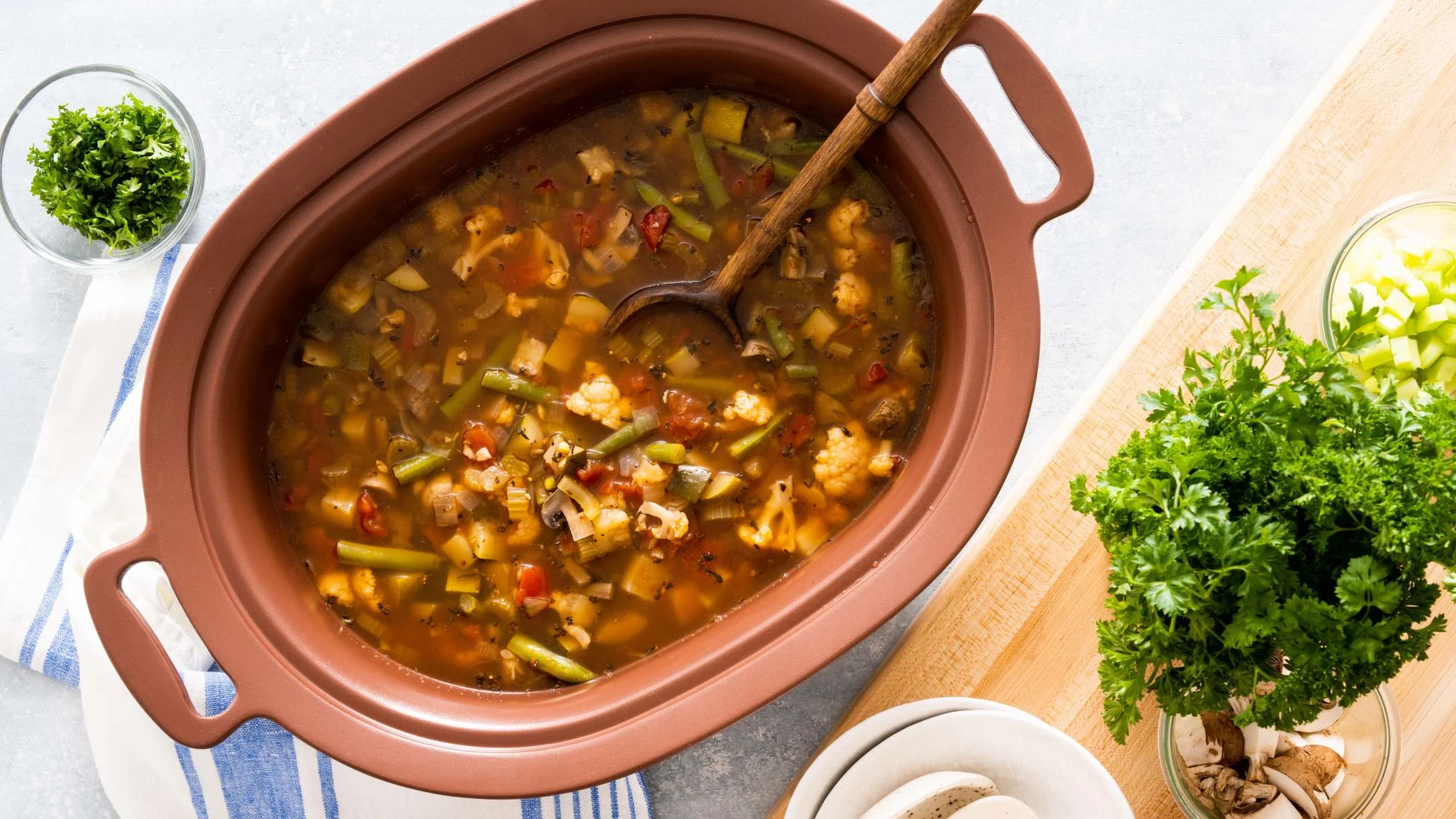 Vegetable soup in slow cooker pot with wooden spoon, surrounded by fresh herbs and ingredients on cutting board.