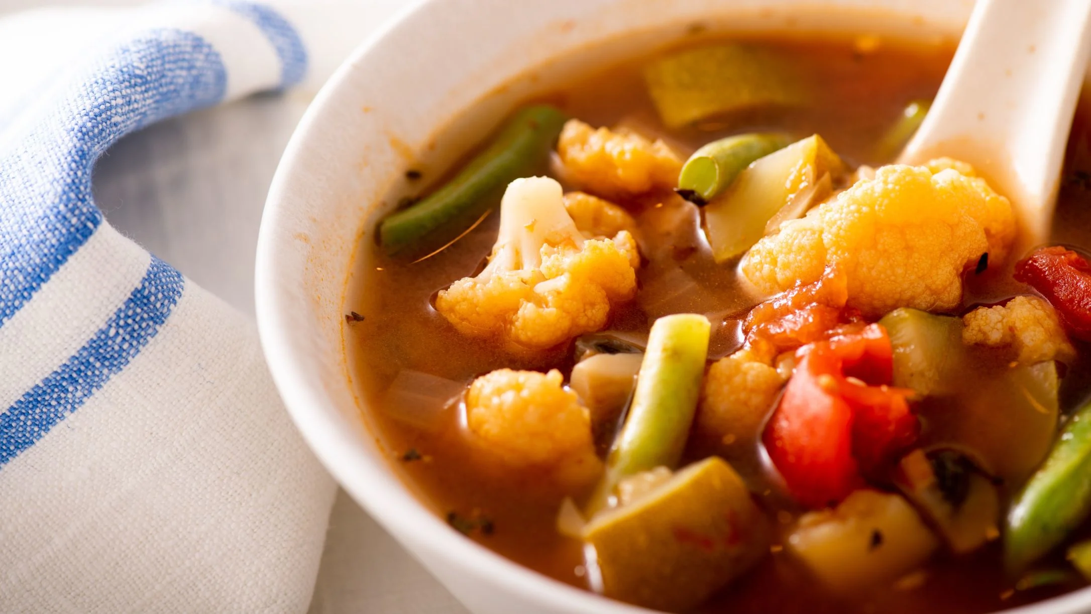 Bowl of vegetable soup with cauliflower, green beans, and tomatoes in broth, served with a blue-striped napkin.