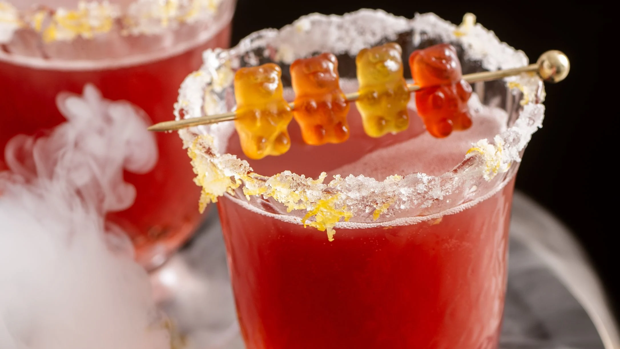 Red cocktail with sugar-rimmed glass garnished with colorful gummy bears on a skewer.