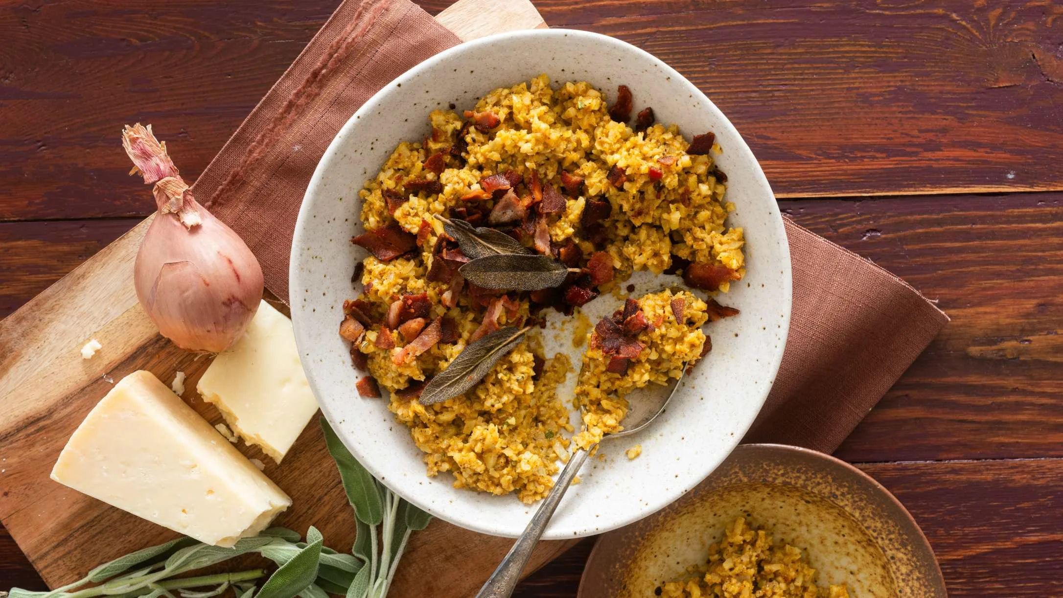 Bowl of yellow risotto with bacon and sage leaves, served with cheese and shallot on wooden cutting board.