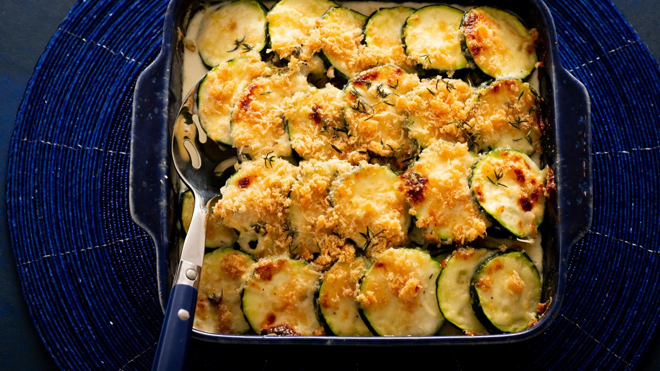 Baked zucchini gratin with golden cheese topping in a blue baking dish with serving spoon.